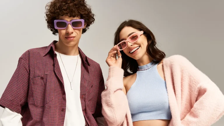 Stylish teen boy with curly hair in purple glasses and burgundy shirt standing next to a brunette girl in pink glasses and fluffy pink jacket, both smiling in a pastel studio setting.