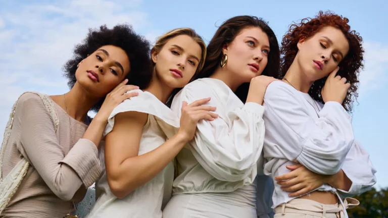A group of four young women with different looks standing close, resting their heads on each other’s shoulders, dressed in soft white and beige tones with the sky in the background