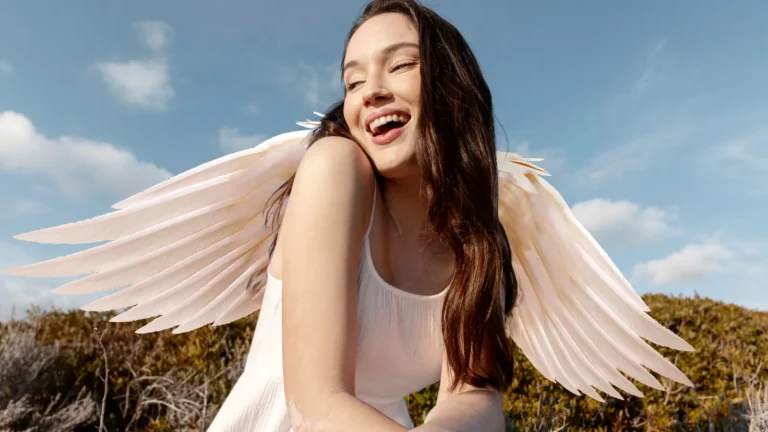 Elegant brunette woman wearing a white flowing dress and angel wings, posing gracefully like a serene angel in a bright setting