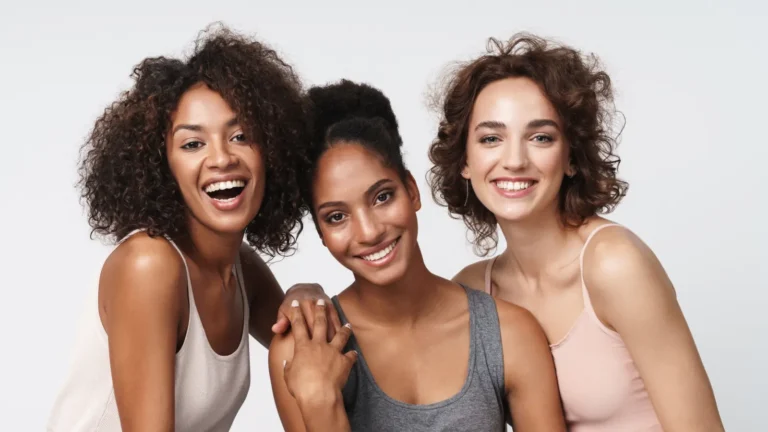 Three beautiful young women smiling and posing together in natural light, showing confidence and elegance in a modern fashion style.